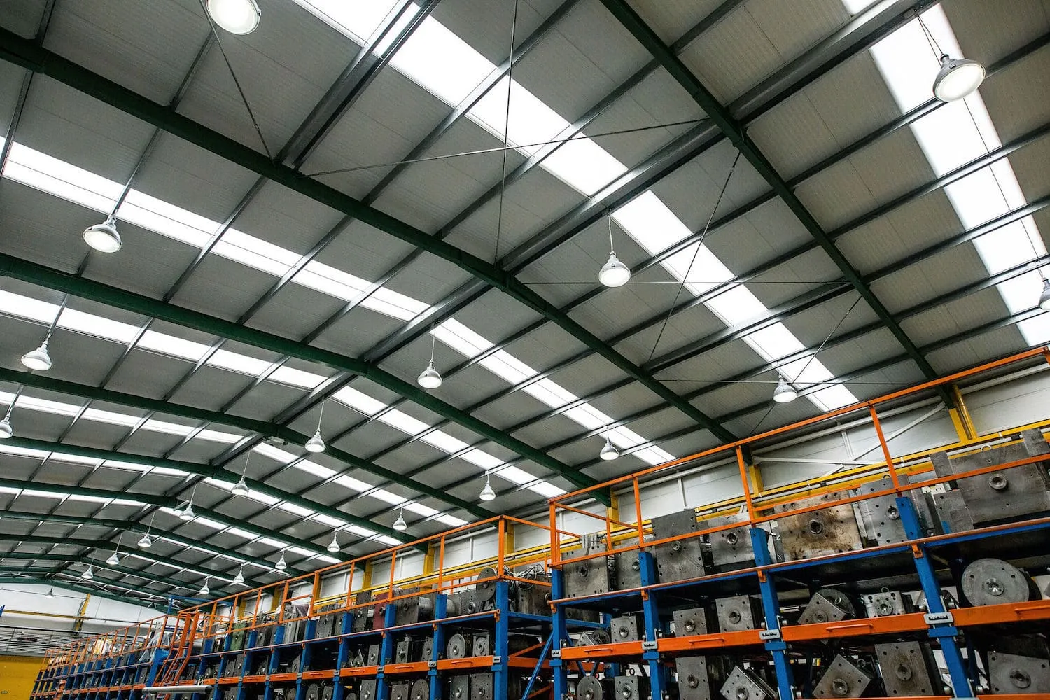 Campanas LED colgadas de la estructura de una nave industrial entre lucernarios, sobre estanterías cargadas